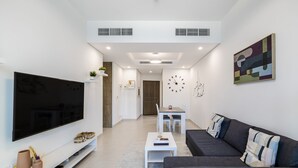 A modern living room with a large flat-screen TV, a dark grey sofa, a white coffee table, and a small dining table with chairs. - The Bay Residences - Canal View (Dubai)
