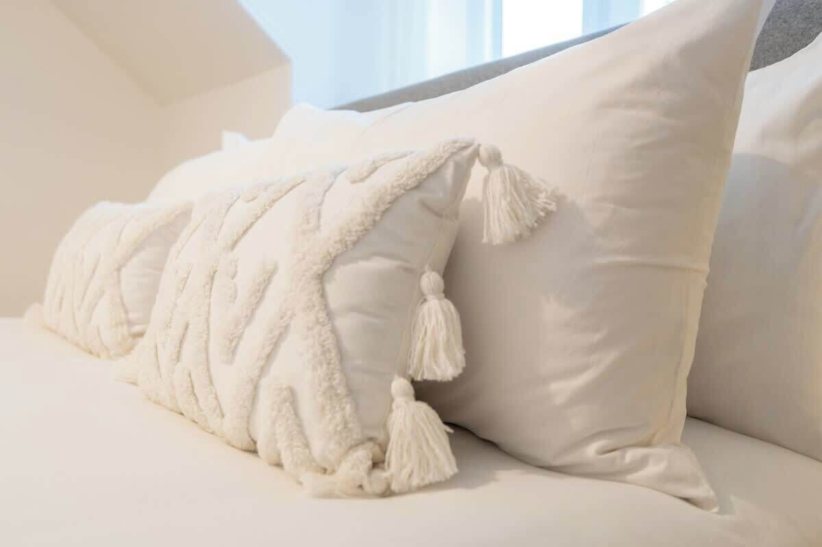 Un lit blanc avec une couverture à motifs et des franges, un oreiller uni et une fenêtre avec des rideaux en arrière-plan.