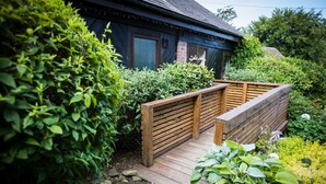 Una terraza de madera que lleva a una casa azul oscuro con una chimenea de ladrillo, rodeada de abundante vegetación.