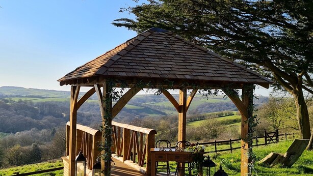 Un pabellón de madera con techo de tejas, situado en una colina cubierta de hierba con vistas a un valle.