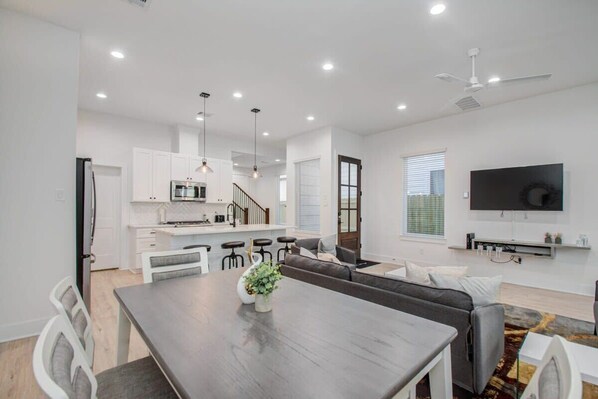 A modern kitchen with white cabinets, a dining area with a dark wooden table, and a living room with a gray sofa and a flat-screen TV. - Modern Farmhouse, Mins Restaurants & Attractions (Houston)