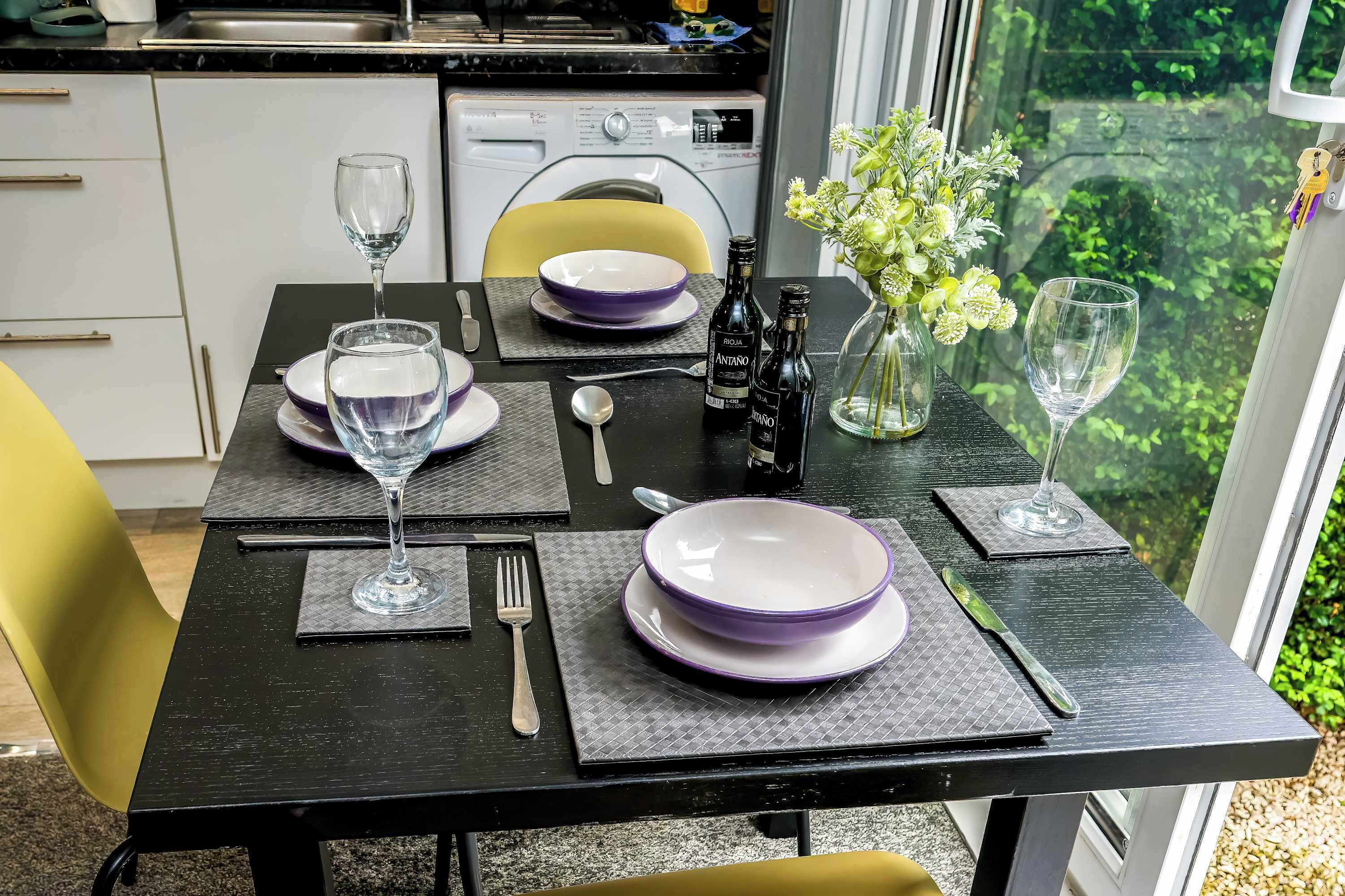 Una mesa de comedor dispuesta con platos morados, copas de vino y una botella de vino Rojalona. Un jarrón con flores está sobre la mesa, y una ventana ofrece vistas de follaje.