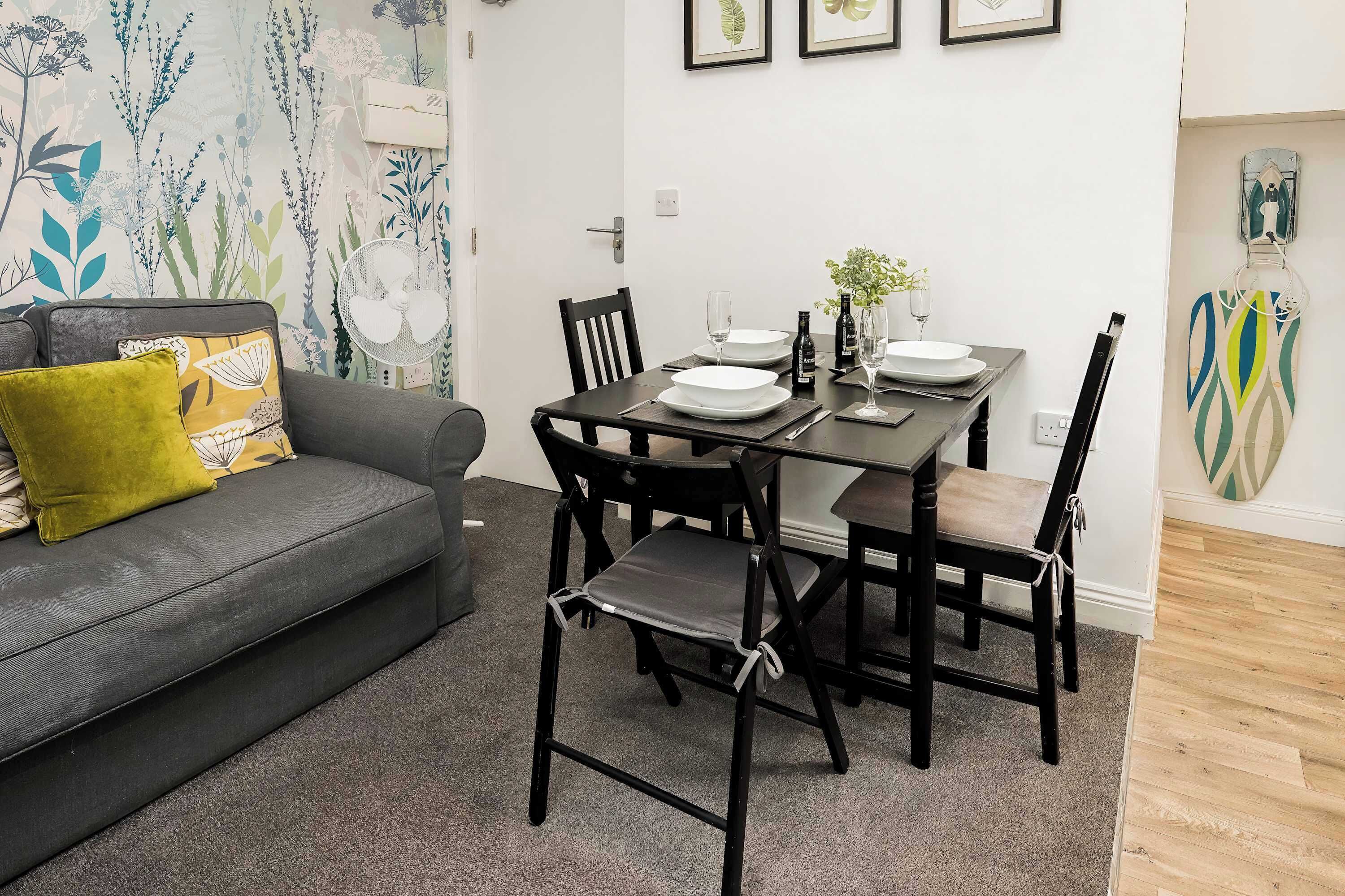 A dining area with a table set for four, surrounded by chairs, a grey sofa with cushions, and a wall with framed artwork.