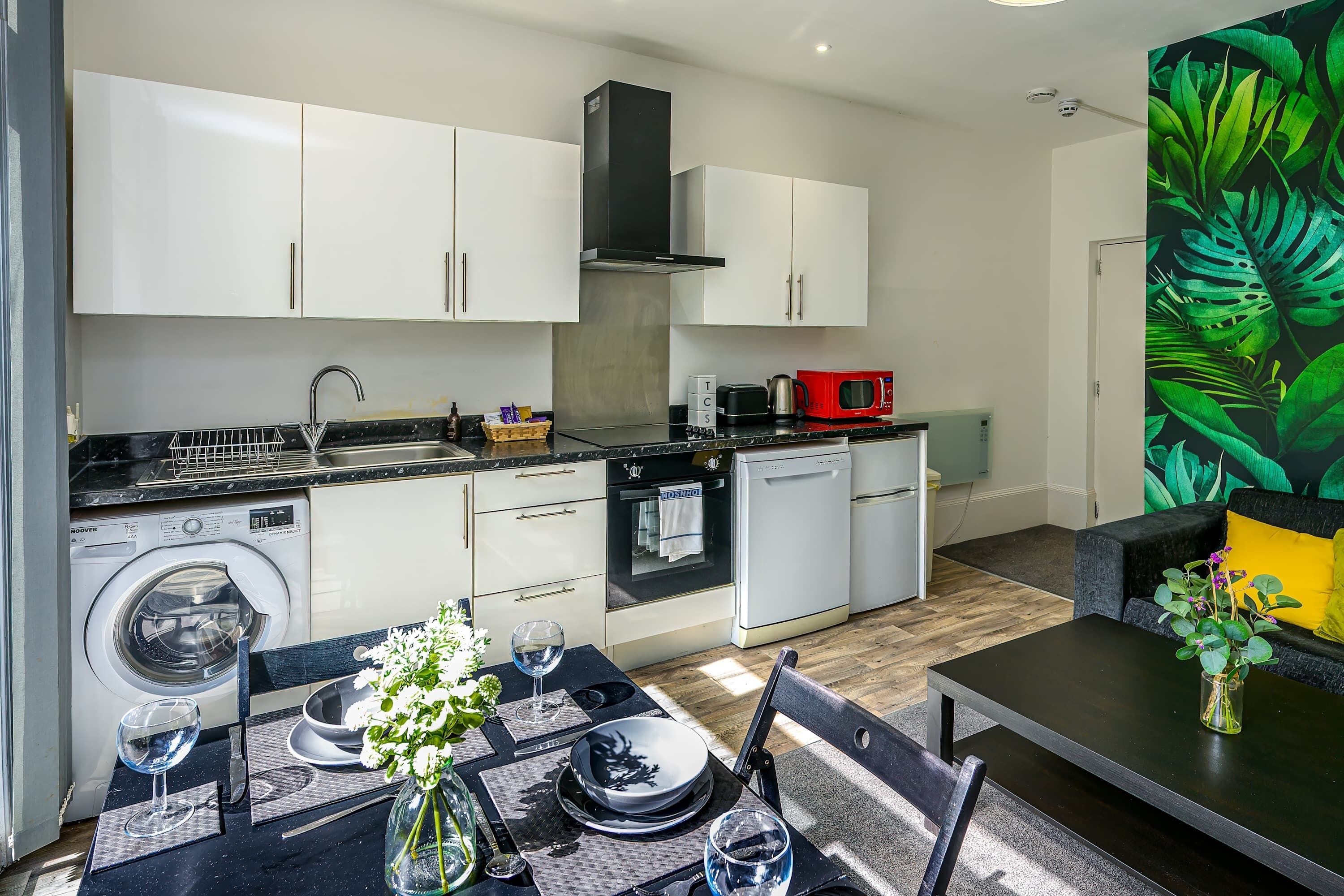 A modern kitchen with white cabinets, a built-in oven, and a microwave. There is a dining area with a table set for two, and a living area with a sofa and a vibrant plant wall.
