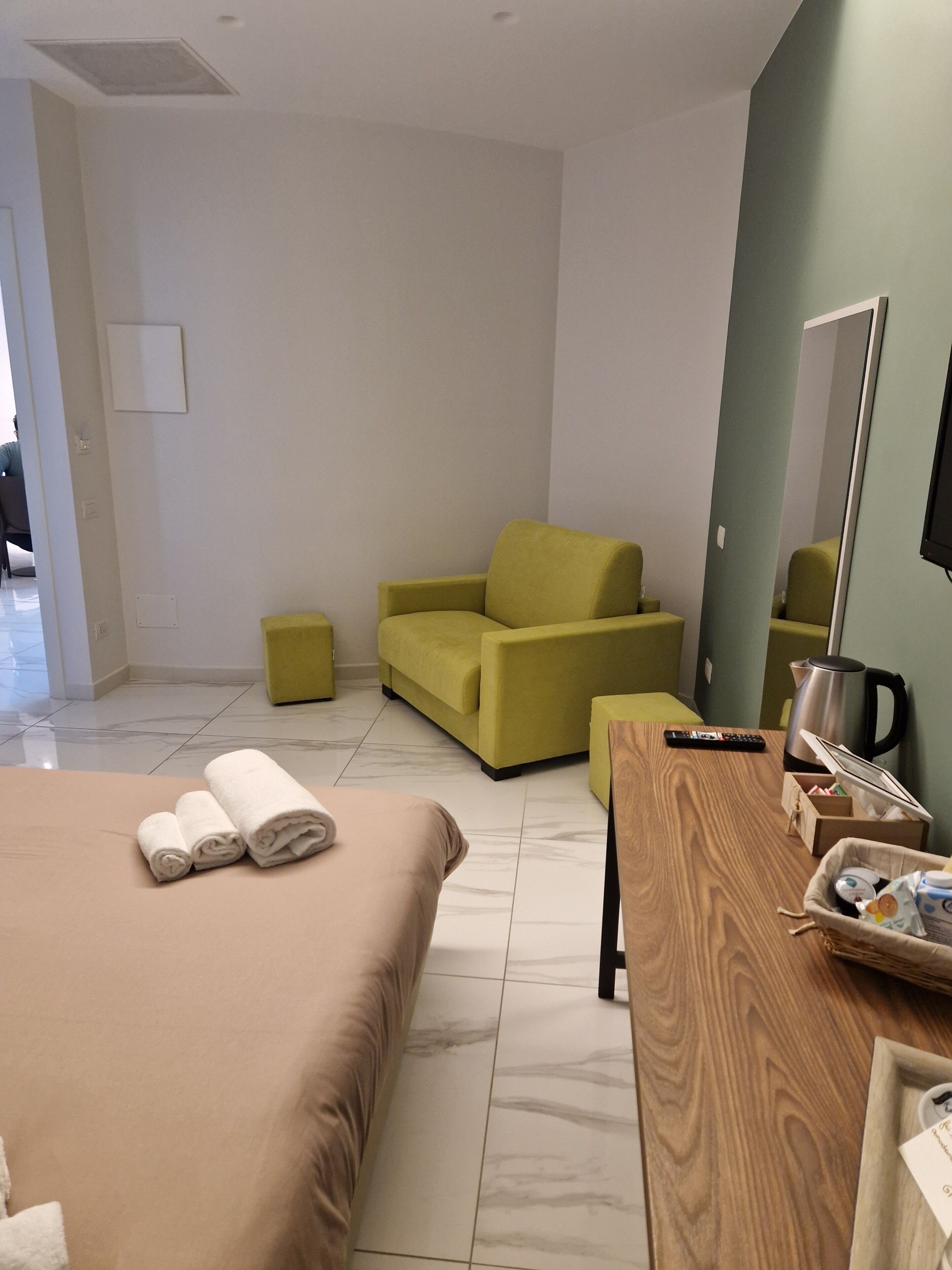 A hotel room with a bed, a wooden table, a yellow armchair, a small stool, a coffee pot, and a remote control.