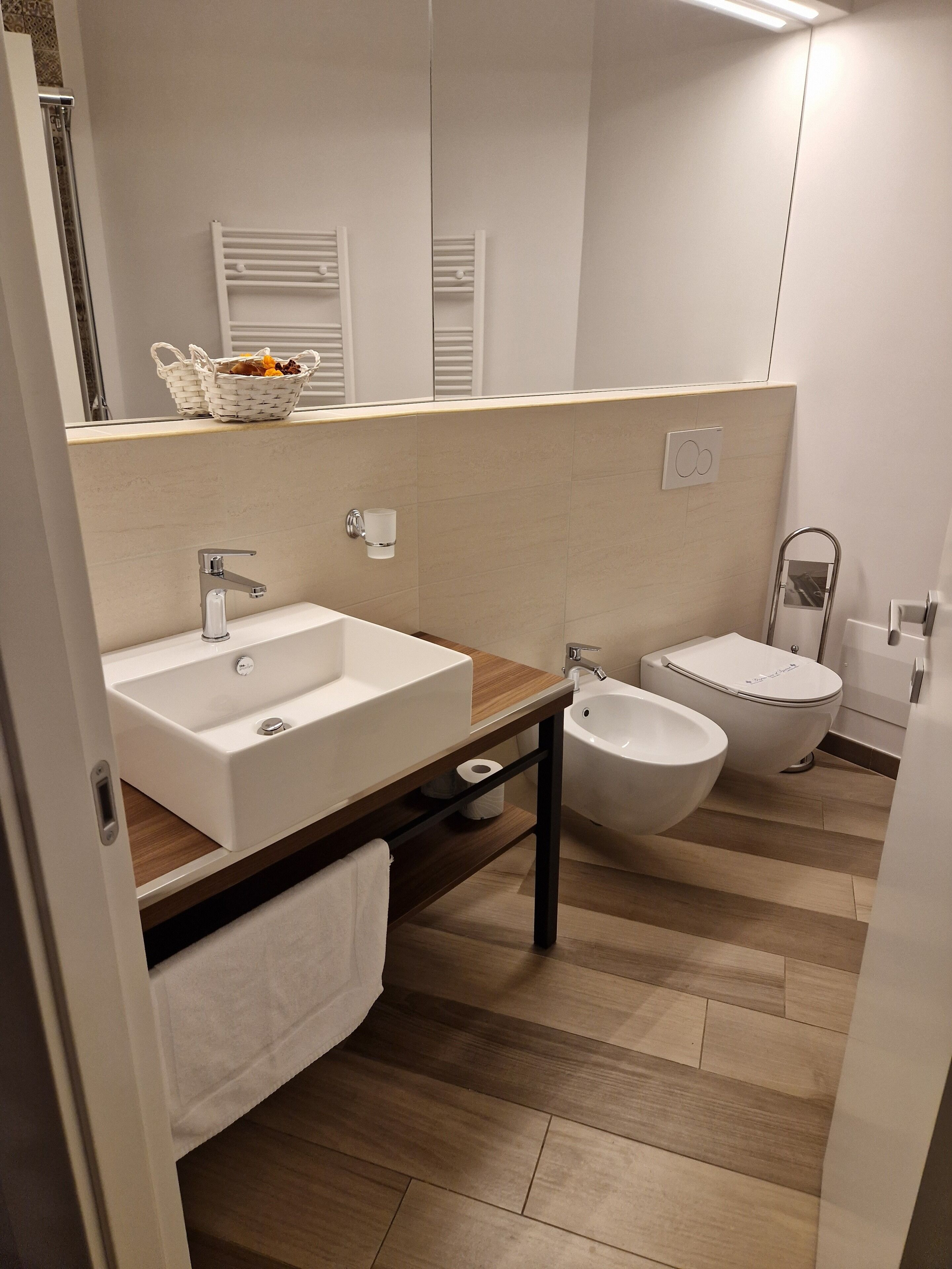 Une salle de bain moderne avec un grand lavabo, un bidet et des toilettes. Un miroir est placé au-dessus du lavabo, et une serviette est suspendue à un porte-serviettes.