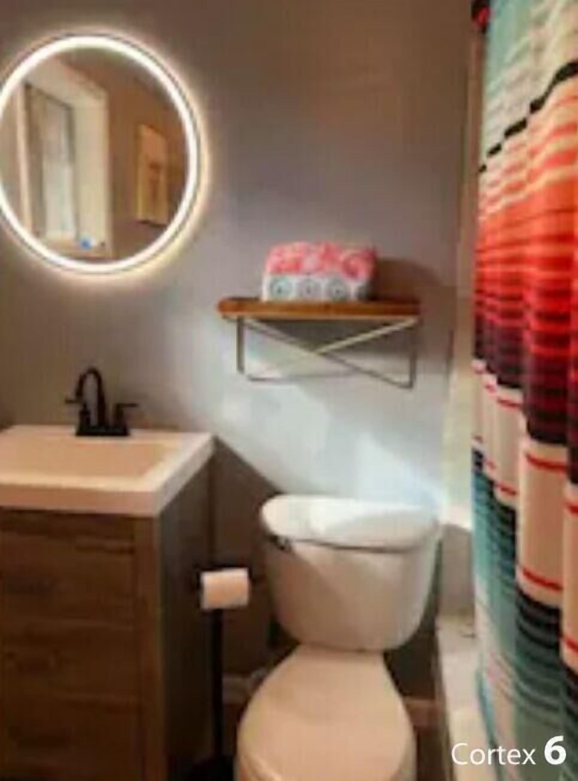 A bathroom with a toilet, a sink, a mirror with a light, and a shelf with towels.