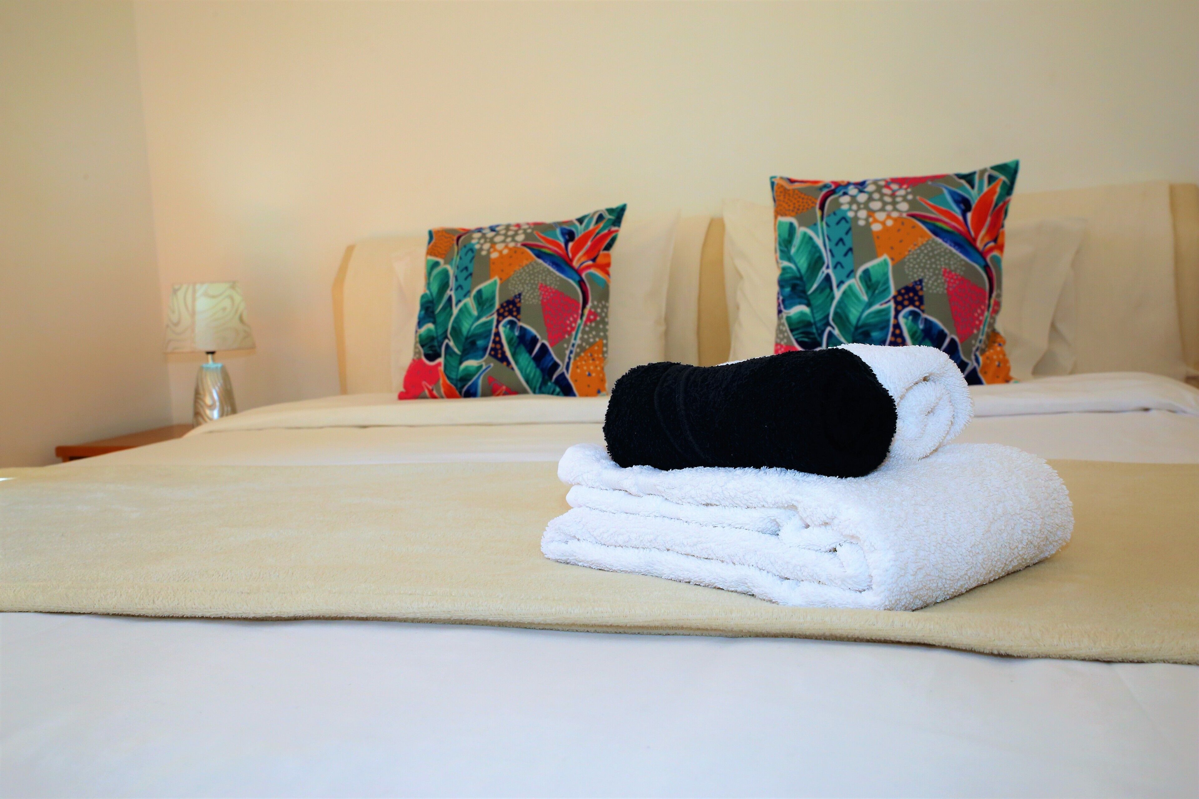 Una cama bien hecha con una toalla blanca doblada y una toalla negra sobre un edredón color beige, flanqueada por almohadas de colores.