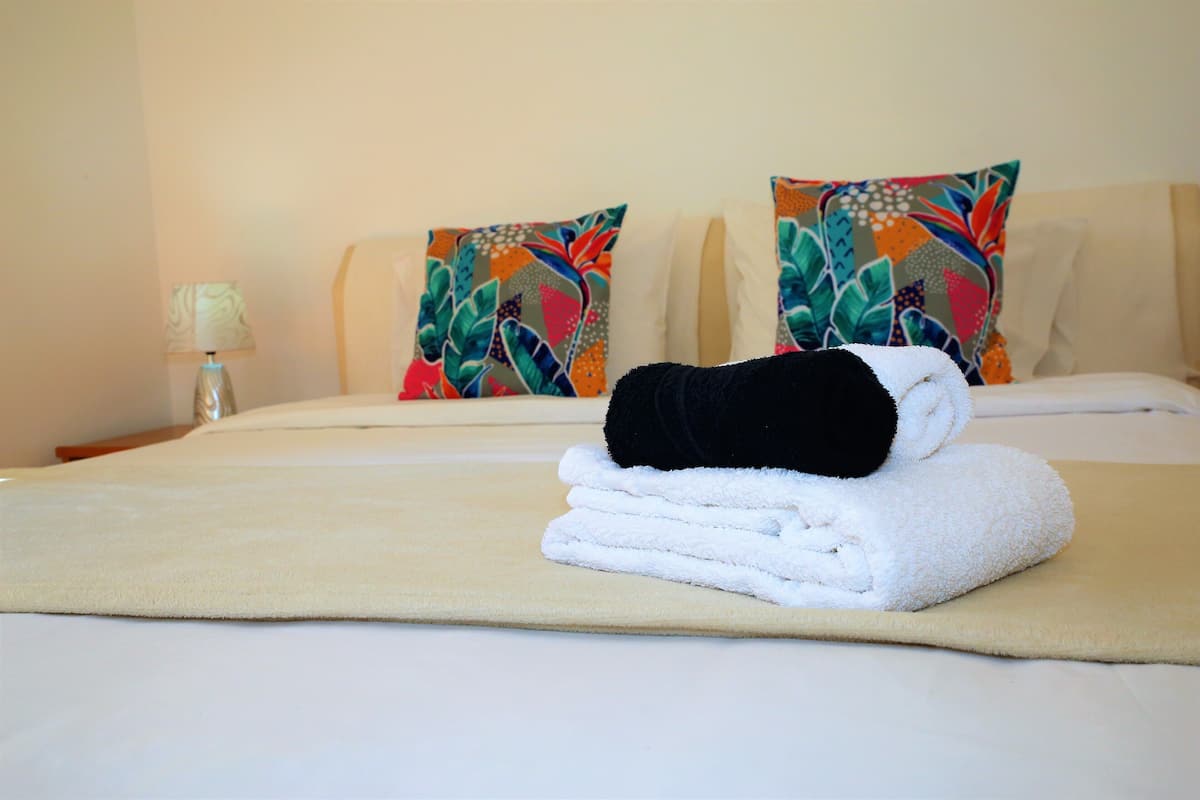 Una cama bien hecha con una toalla blanca doblada y una toalla negra sobre un edredón color beige, flanqueada por almohadas de colores.