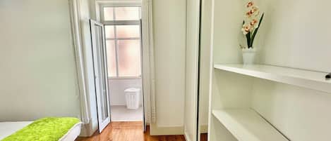 A narrow room with a wooden floor, a bed with a green blanket, a white shelf with flowers, and a view through a door to a balcony.