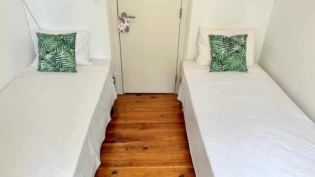 Una habitación con dos camas individuales, cada una con un colchón blanco y una almohada estampada con hojas verdes. En el centro, una puerta de madera, y las paredes son blancas.