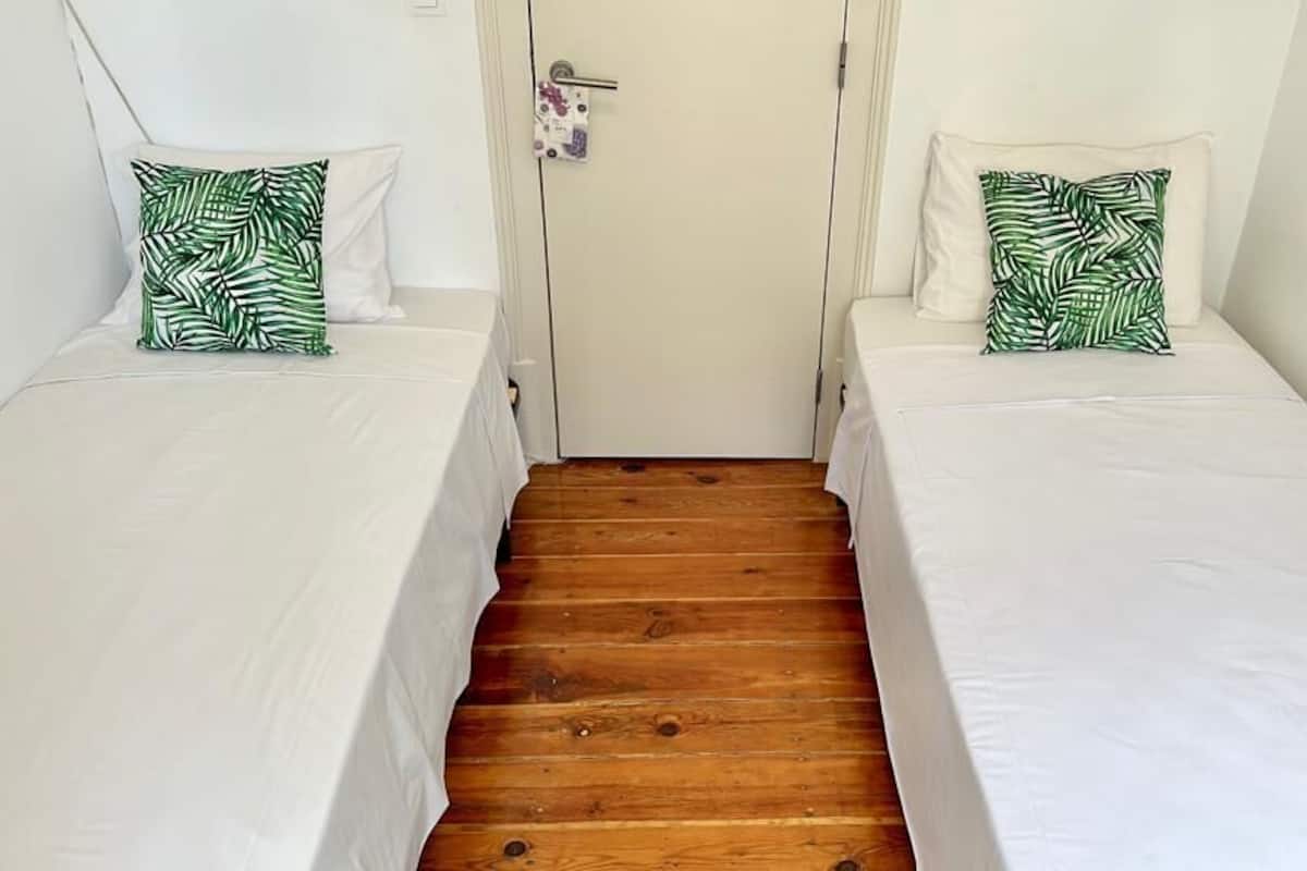 Una habitación con dos camas individuales, cada una con un colchón blanco y una almohada estampada con hojas verdes. En el centro, una puerta de madera, y las paredes son blancas.