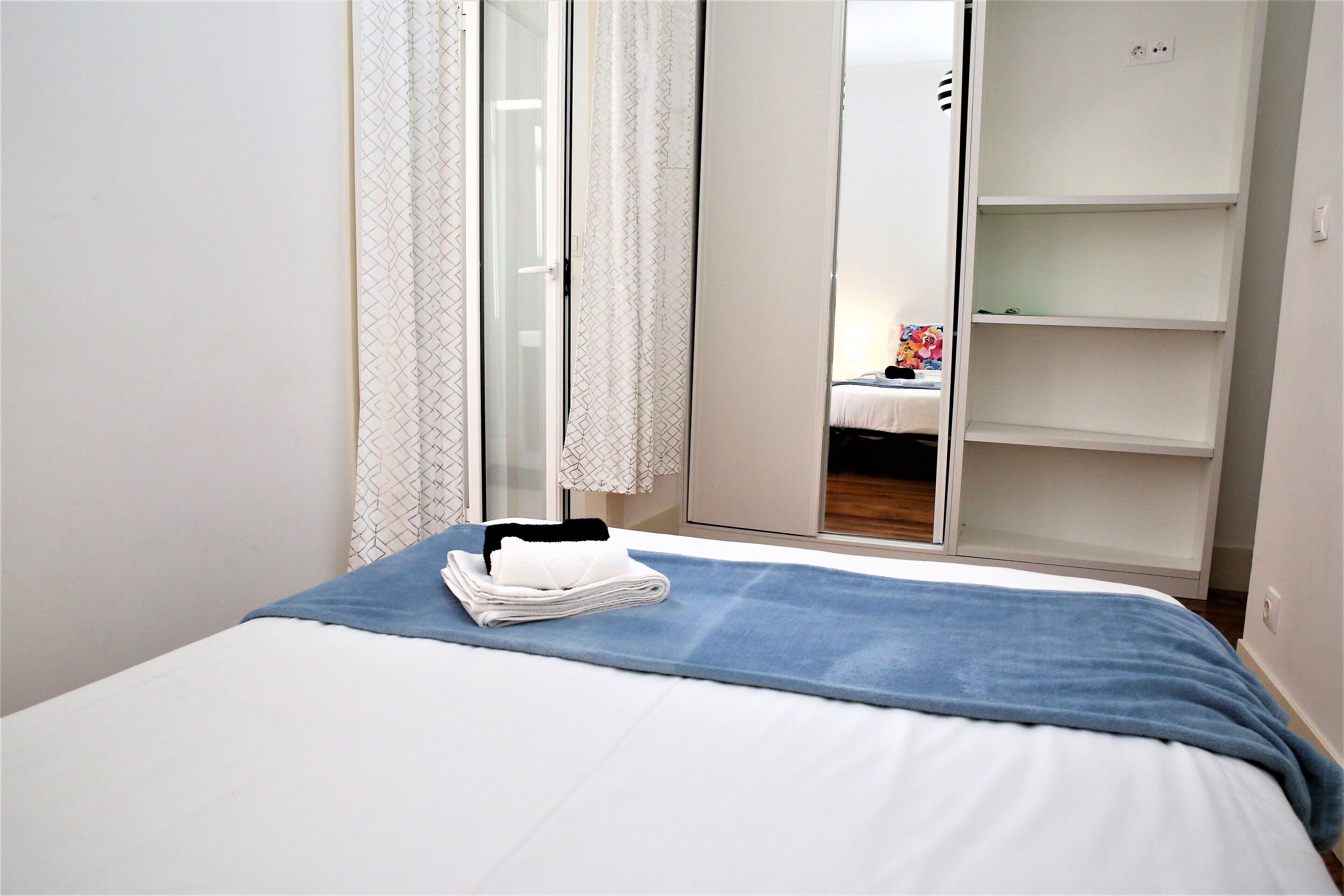 Una cama bien hecha con una colcha azul y toallas blancas dobladas, un armario blanco con las puertas abiertas y un espejo que refleja un objeto colorido sobre una mesa.