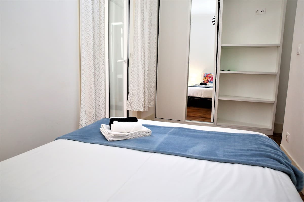 Una cama bien hecha con una colcha azul y toallas blancas dobladas, un armario blanco con las puertas abiertas y un espejo que refleja un objeto colorido sobre una mesa.