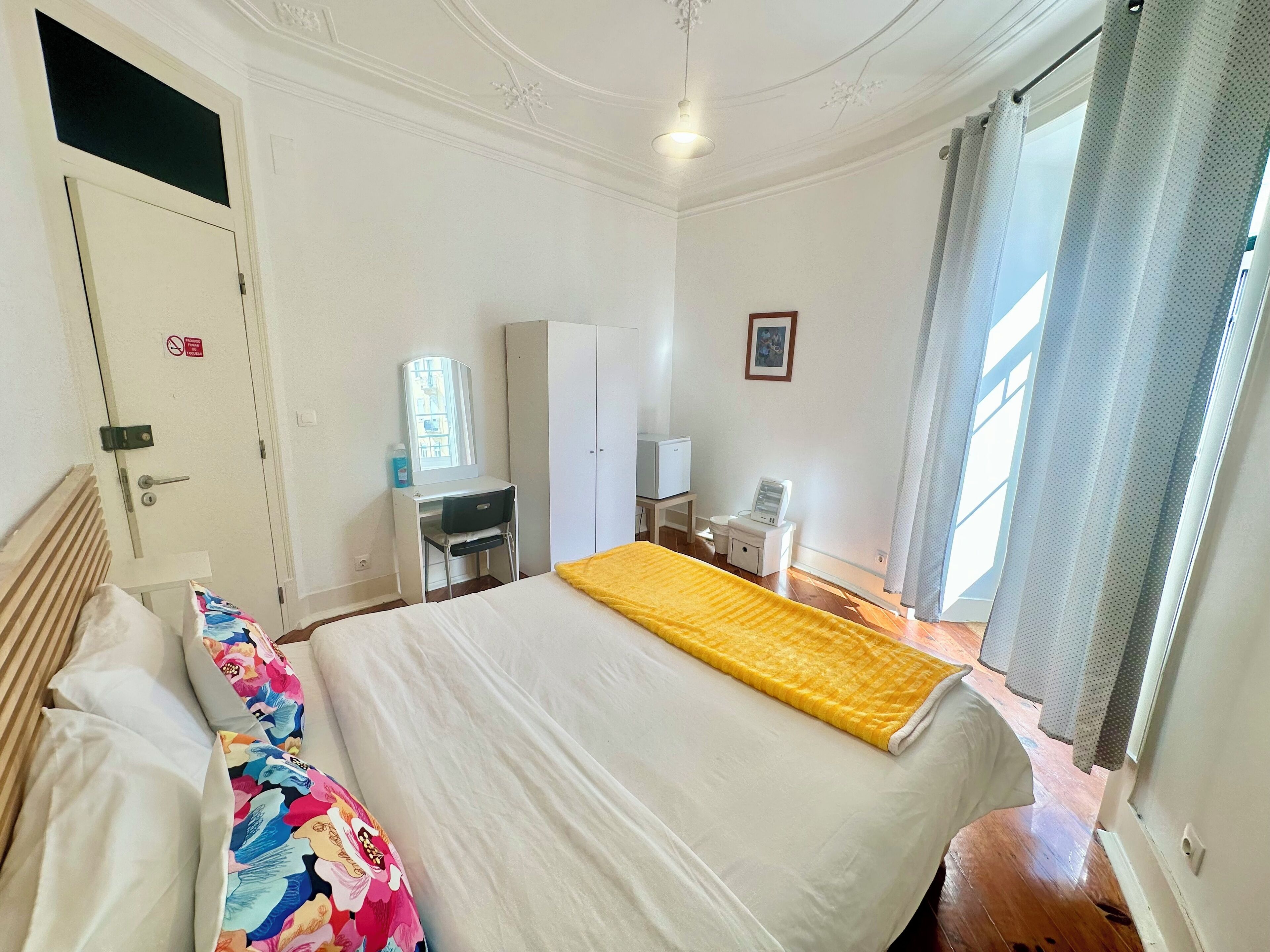 Habitación de hotel con cama, escritorio, armario, mesita con lámpara y ventana con cortinas.