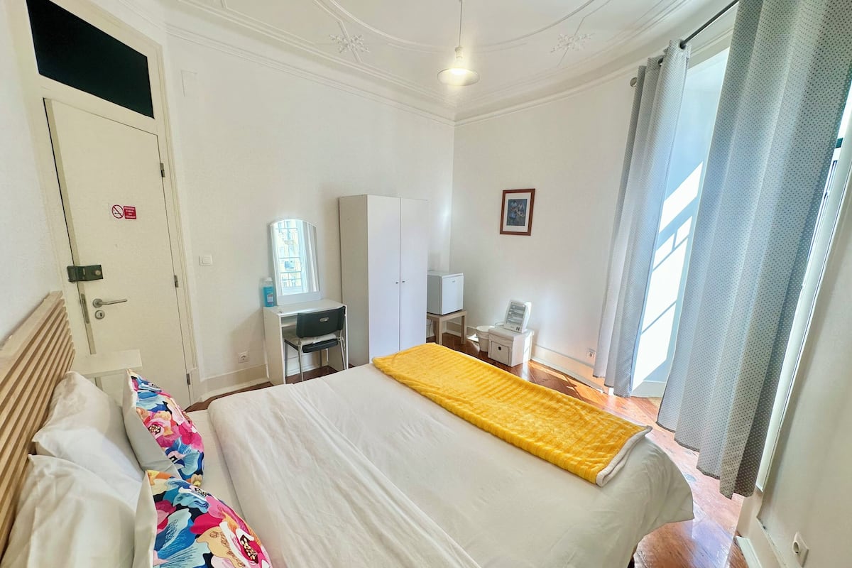 Habitación de hotel con cama, escritorio, armario, mesita con lámpara y ventana con cortinas.