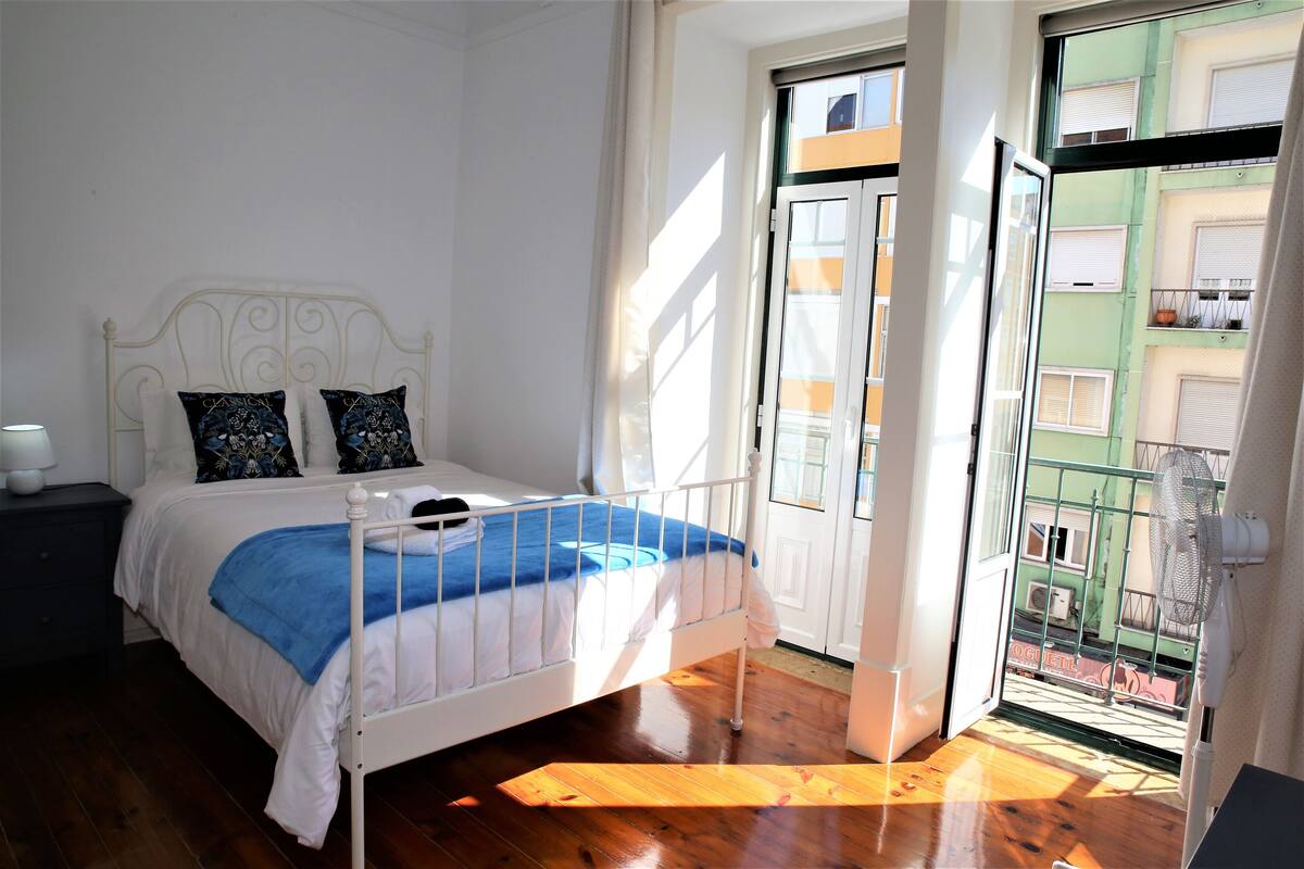 Un dormitorio con una cama, una mesita de noche, una ventana con vistas a edificios y una puerta.