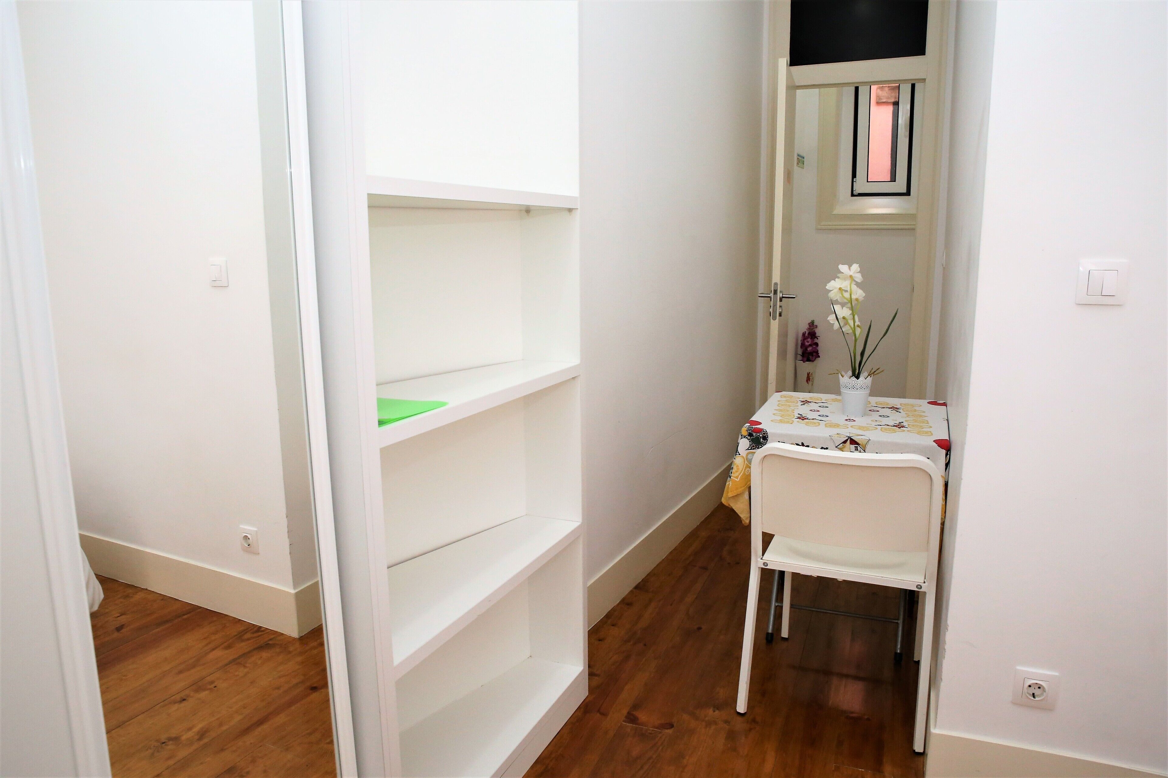 A narrow hallway with white walls and wooden flooring, leading to a small room with a white chair and a table with a vase of flowers.