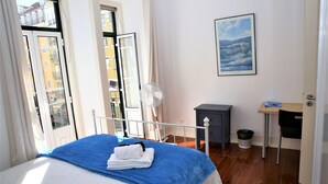 Una camera da letto con un letto, una ventola da terra, un tavolino con una sedia e un quadro appeso al muro.