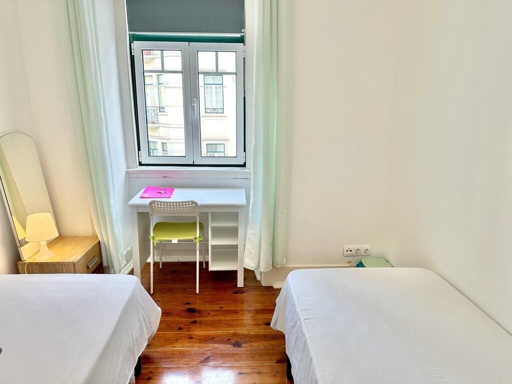 Une petite chambre propre, avec deux lits simples, un bureau avec une chaise, une fenêtre avec des rideaux et une lampe.