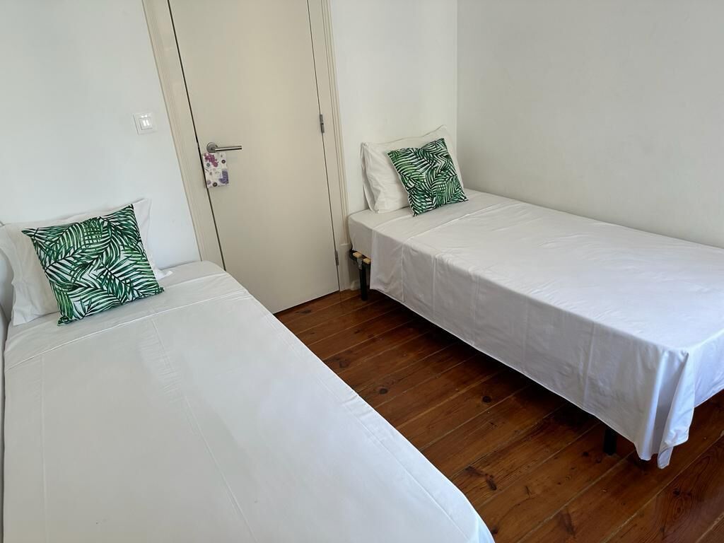 Zwei Einzelbetten mit weißer Bettwäsche und Kissen mit grünem Blattmuster in einem Zimmer mit Holzboden und weißen Wänden.