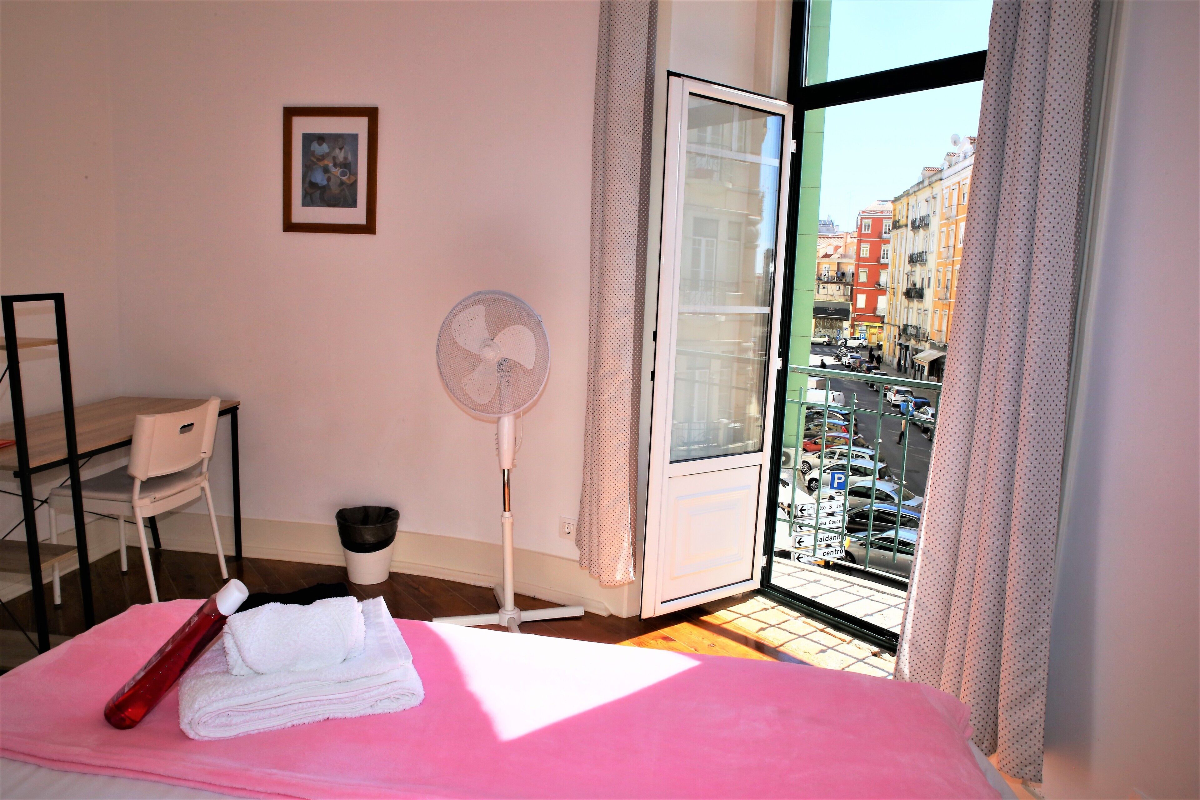 A hotel room with a bed, a fan, a chair, and a view of the city through a large window.