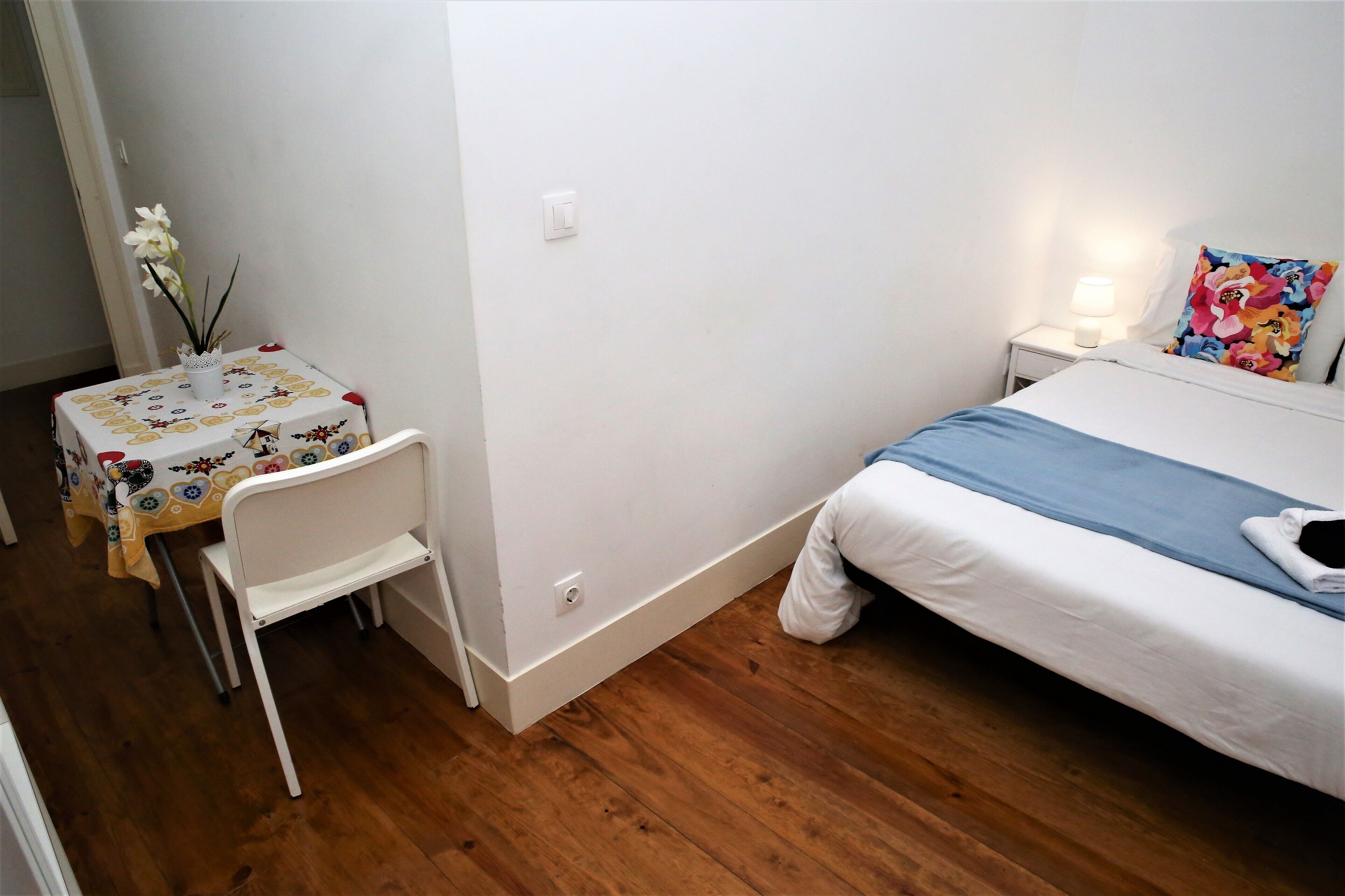 Una camera da letto con un letto, un comodino, una lampada, un tavolino con una sedia e un vaso di fiori.