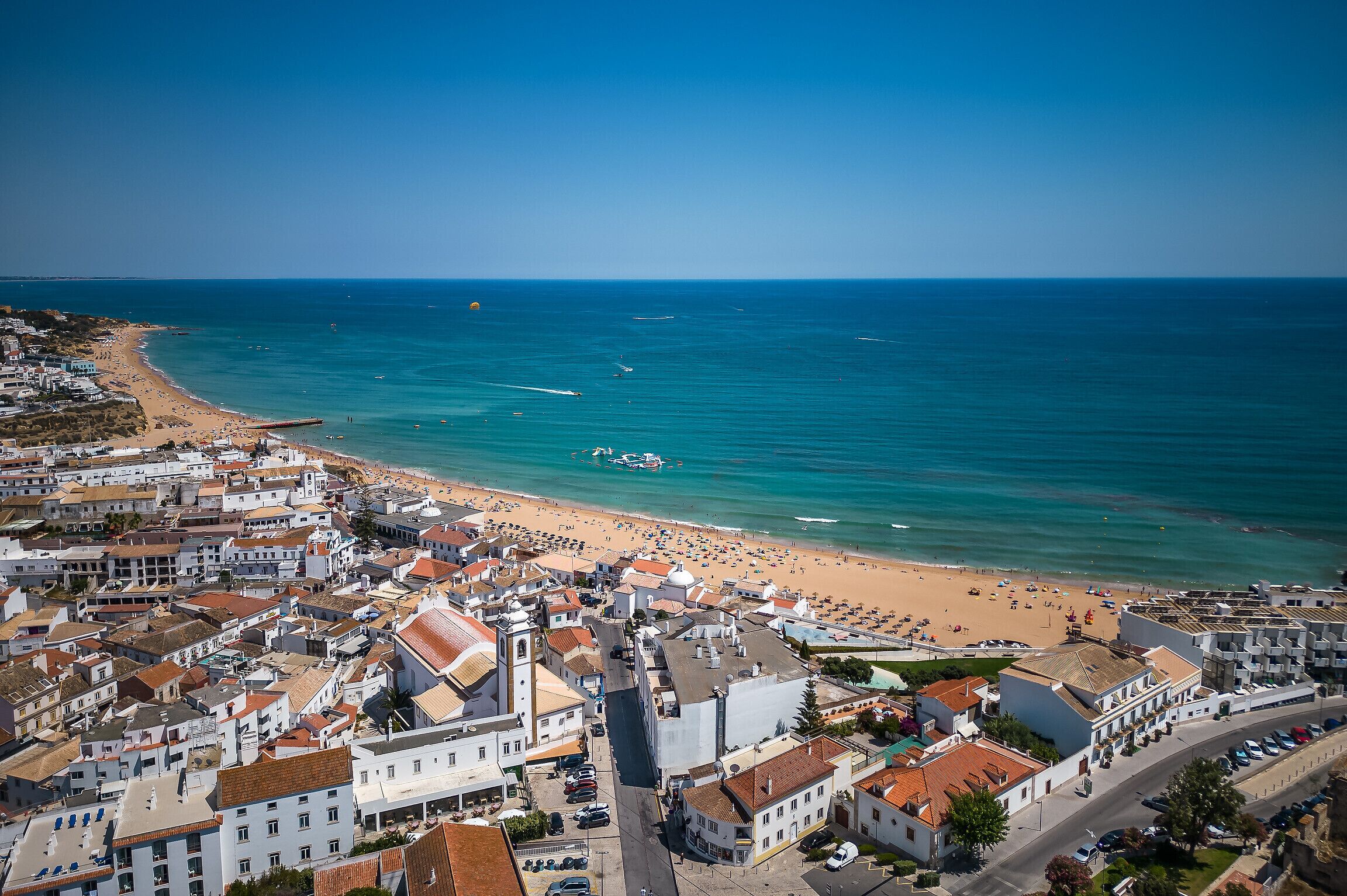An aerial view of a coastal town with a beach, buildings, and clear blue waters.