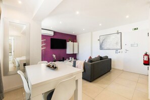 A modern living room with a white dining table, grey sofa, and a flat-screen TV mounted on the wall.