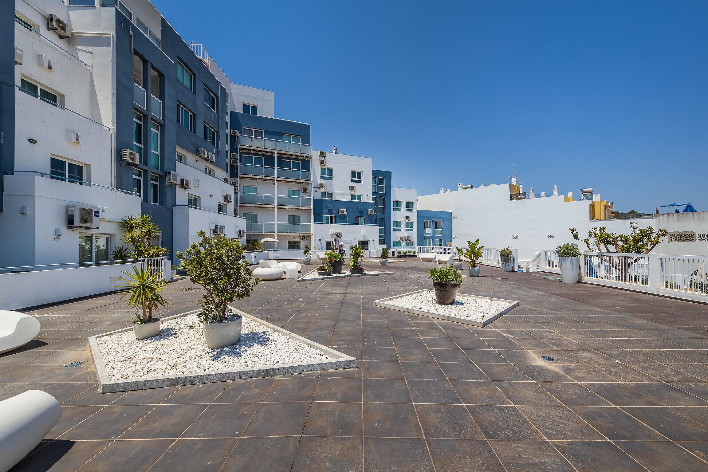 Eine Dachterrasse mit Blumentöpfen, Sitzbereichen und blauem Himmel.