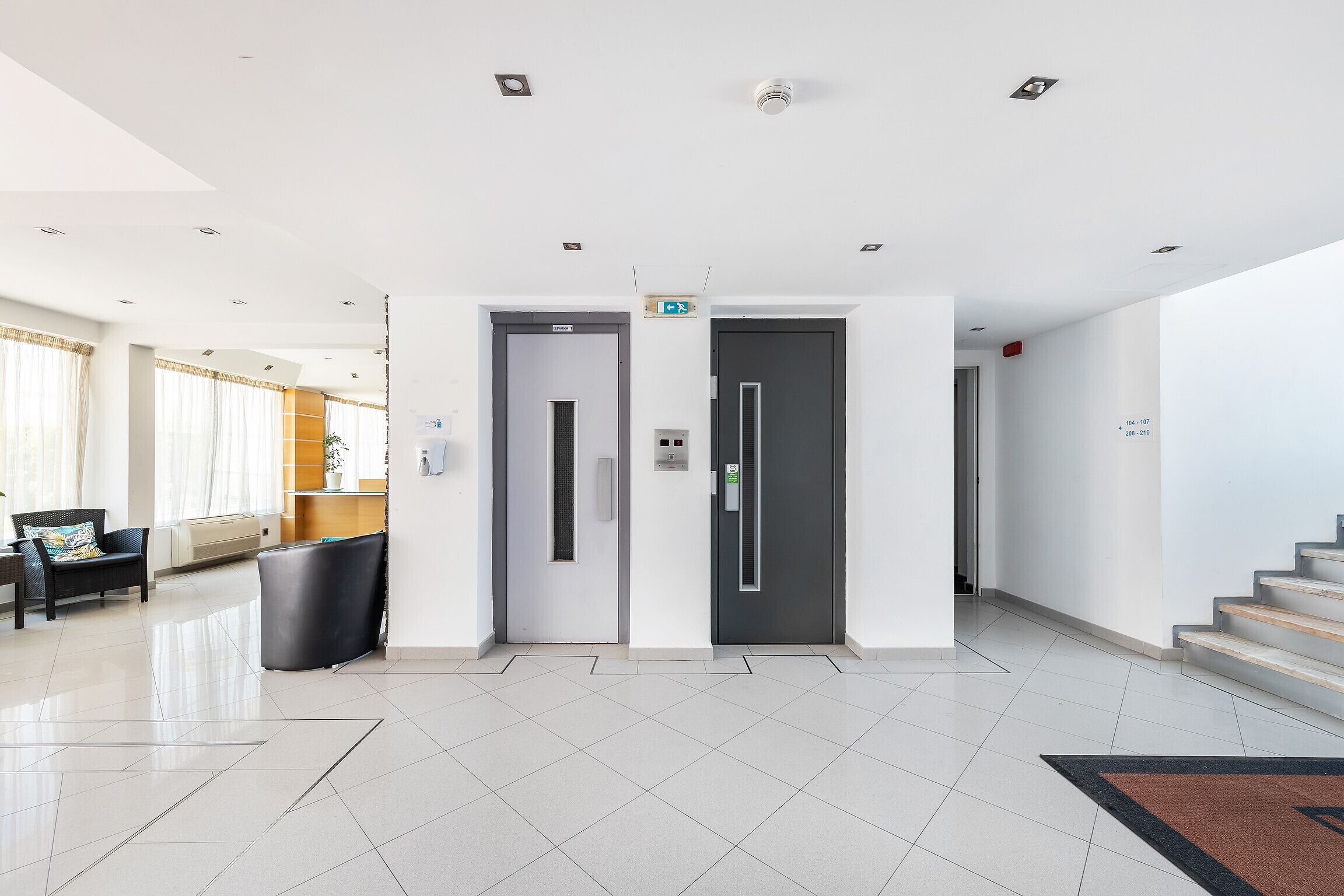 A modern lobby with a reception desk, two elevator doors, and a staircase.