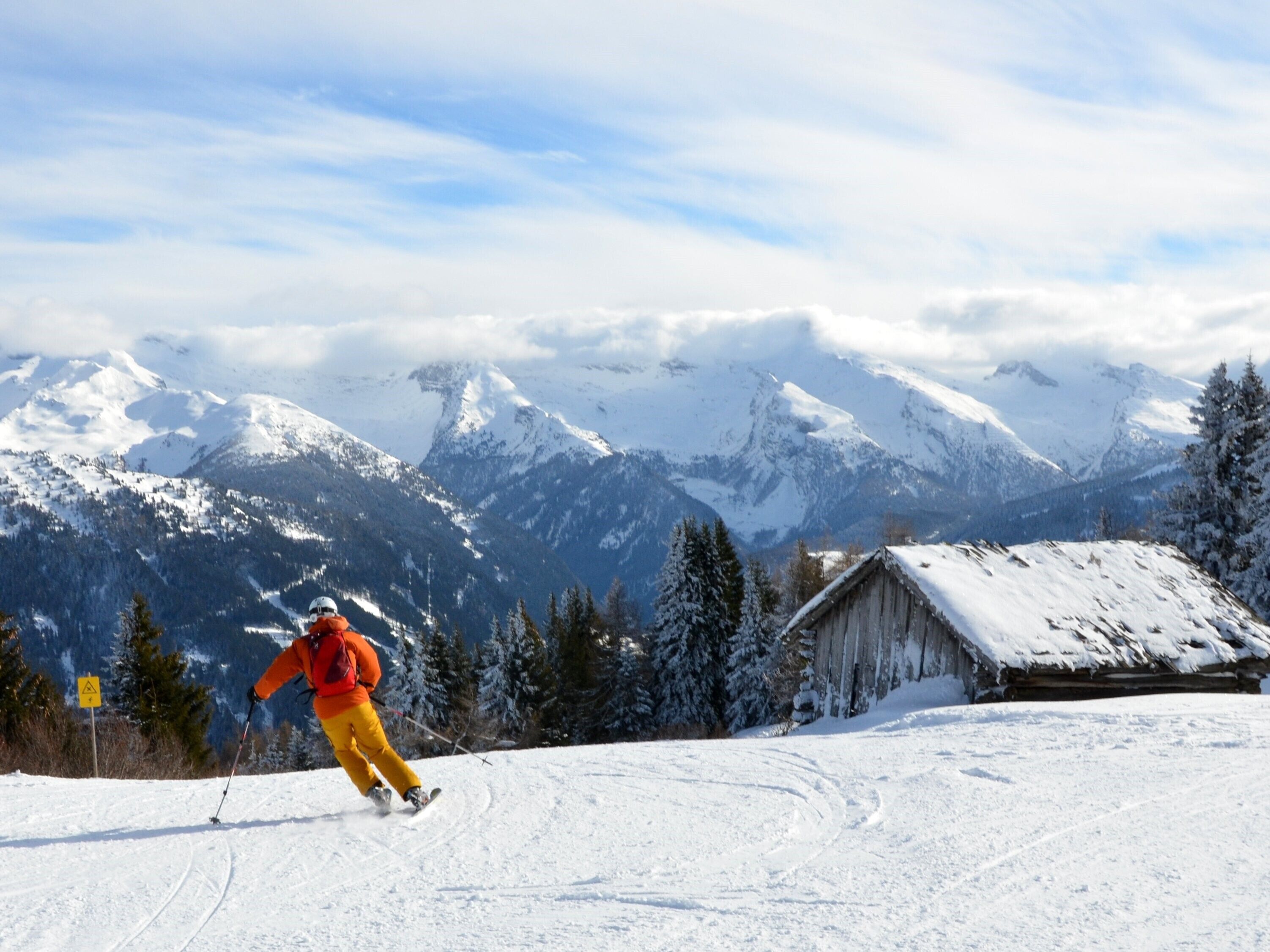 Un paysage de montagne enneigé avec un skieur en mouvement, un chalet en bois et des arbres recouverts de neige.