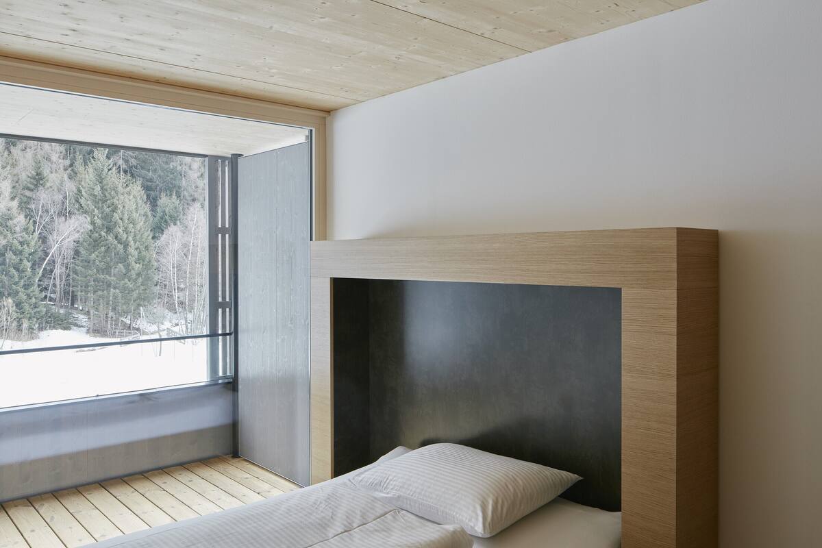 Un dormitorio moderno con techo de madera, un ventanal con vistas a un bosque y una cama con dos almohadas.