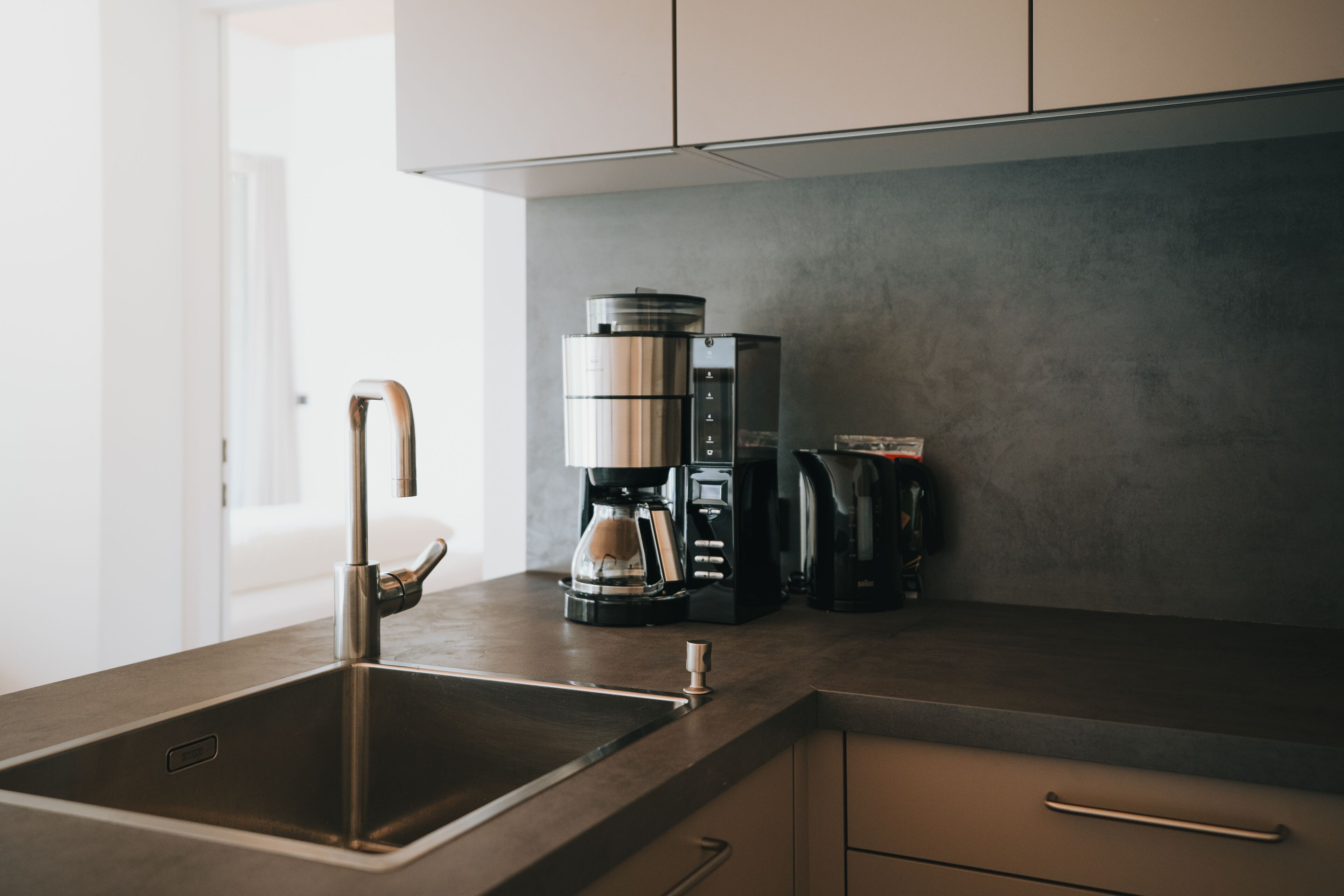 A modern kitchen with a stainless steel sink, a coffee maker, and a kettle on the countertop.