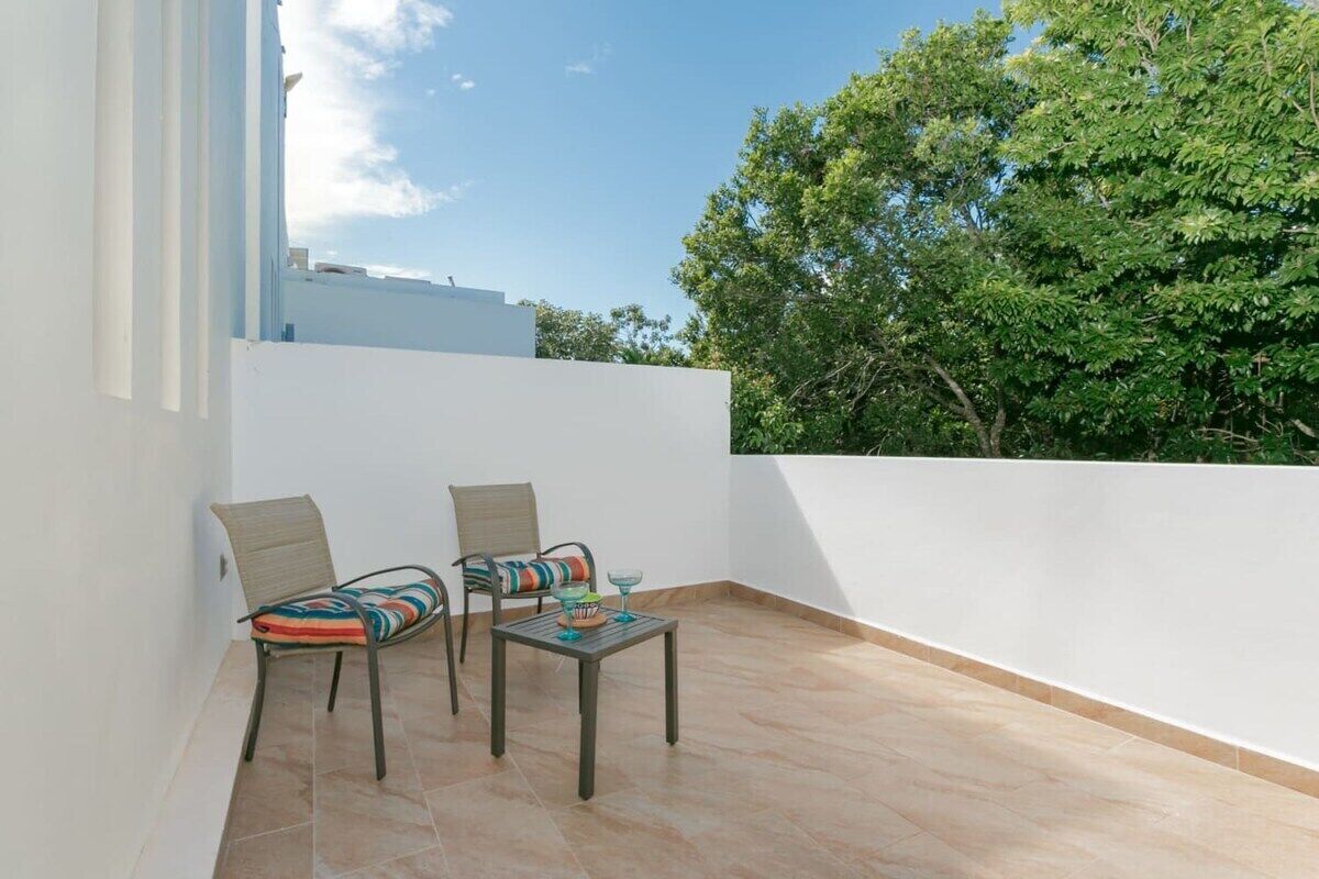A balcony with two chairs and a small table, white walls, and a tree in the background.
