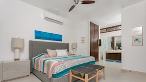 Ein Schlafzimmer mit Bett, Nachttischlampen, Deckenventilator, Fenster und einem im Hintergrund sichtbaren Badezimmer.