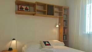 Un lit bien fait, avec du linge de lit blanc et un oreiller orné d’un motif de voilier, une lampe de chevet, une table de chevet en bois et une bibliothèque remplie de livres et d’un tableau encadré.