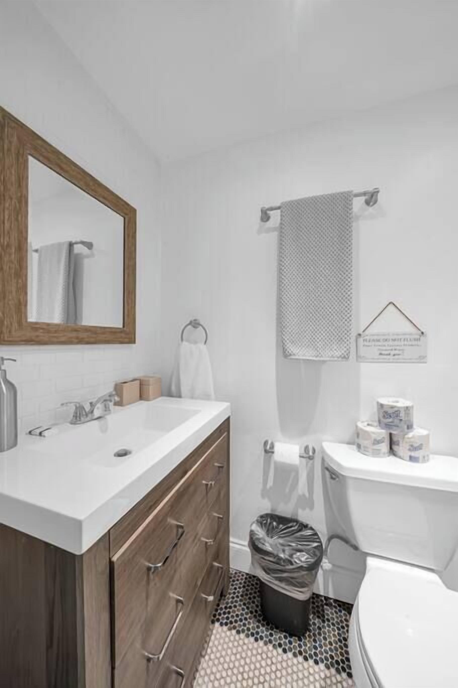 Ein Badezimmer mit einem weißen Waschbecken, einem Holz-Badmöbel, einem Spiegel, einem Handtuchhalter, einer Toilette und einer schwarzen Mülltonne.