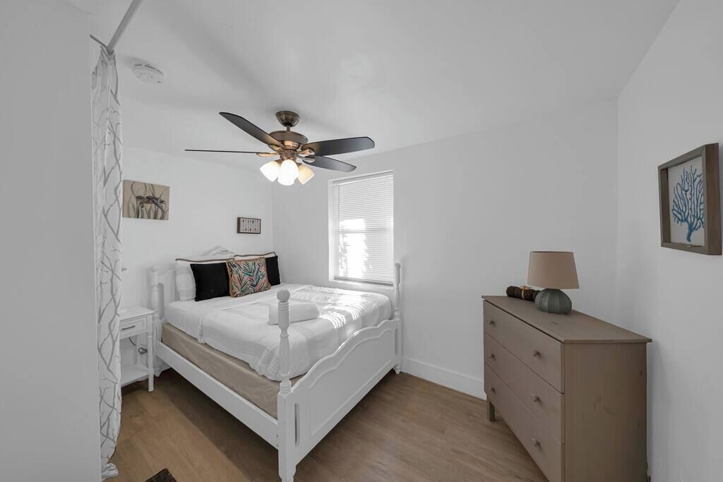 A bedroom with a white bed, a ceiling fan, a dresser, and a window with blinds.
