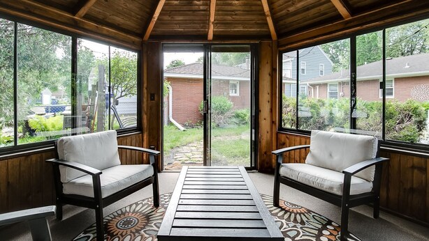 A covered patio with two chairs and a patterned rug, large windows, and a view of a backyard. - Charming Mid-Century Modern Eco-Friendly Home (Saint Paul)