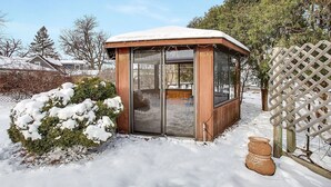 A small, enclosed wooden structure with a glass door, surrounded by snow-covered ground and trees. - Charming Mid-Century Modern Eco-Friendly Home (Saint Paul)
