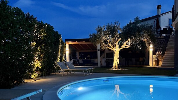 A swimming pool with a curved edge, surrounded by a paved deck. There are lounge chairs and a covered seating area with a tree in the center. - Romantic vacation apartment with large pool and beautiful garden (Bale)