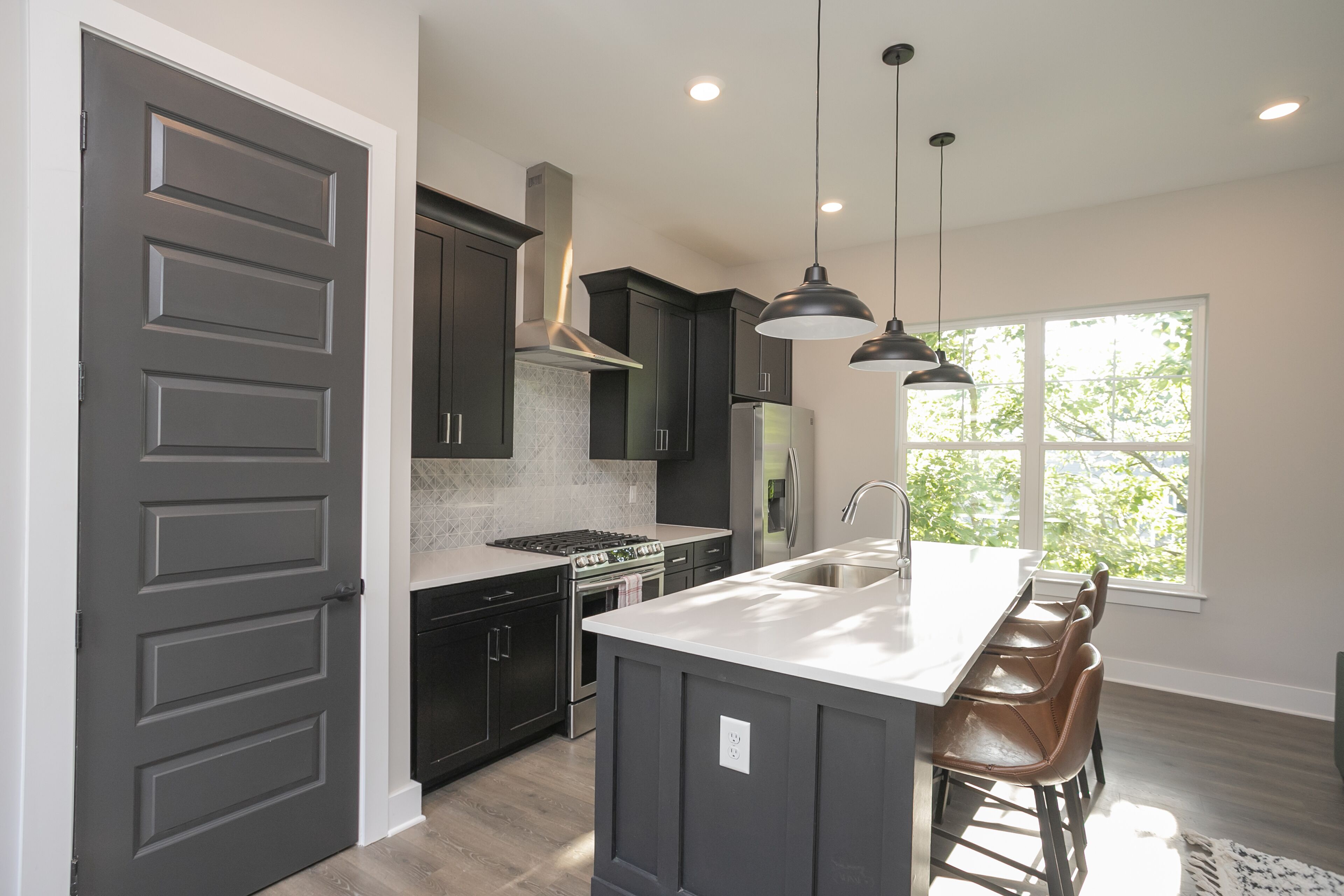 A modern kitchen with dark cabinetry, a central island with bar stools, a large window with a view of trees, and pendant lighting.