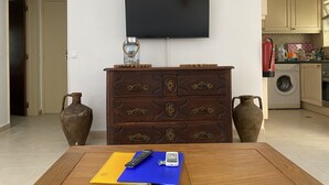 A living room with a wooden table, a TV mounted on the wall, a dresser with drawers, and a kitchen area visible through an archway. - Carvoeiro Sol Right by the sea (Carvoeiro)