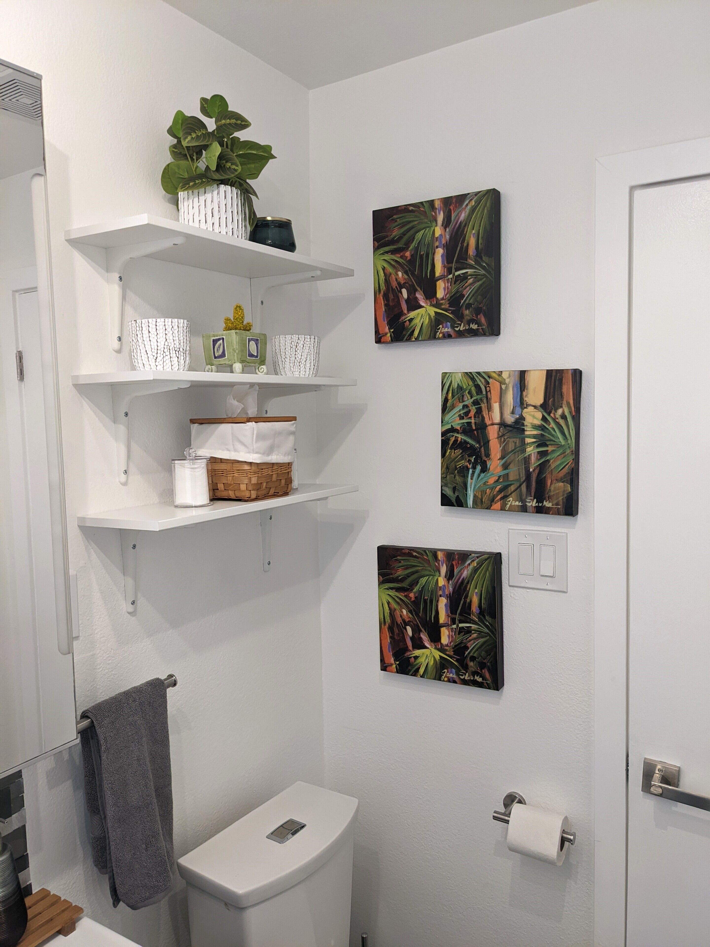 Um banheiro com vaso sanitário, prateleiras com diversos objetos e três quadros decorativos na parede.