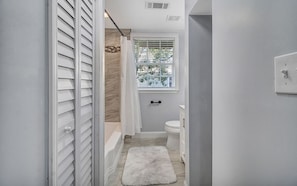 A narrow bathroom with a bathtub, toilet, and window with blinds.