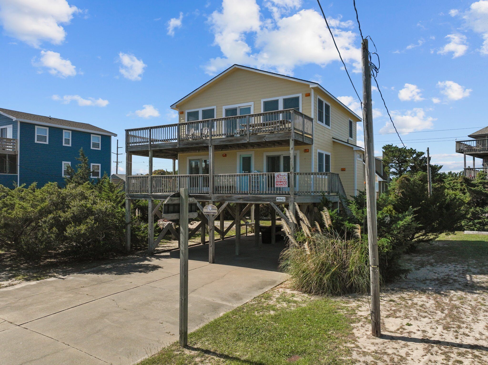 A two-story beachfront house with a balcony, a 'For Sale' sign, and a sandy beach in the background.