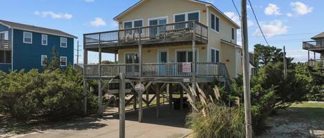 A two-story beachfront house with a balcony, a 'For Sale' sign, and a sandy beach in the background.