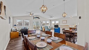 A spacious dining area with a wooden table set for four, surrounded by chairs. There are ceiling fans, a large fish decoration, and a view of the outside through large windows.