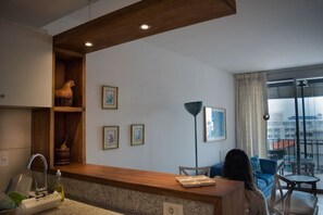 A modern living room with a wooden reception desk, a sofa, and a dining table. There are framed pictures on the wall and a large window with a view of buildings.